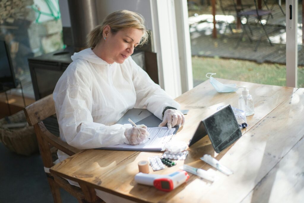 A healthcare worker in protective gear documents patient data indoors during daylight.