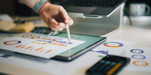 Hand using a tablet and charts on desk to analyze business data. Ideal for finance, business, and technology themes.