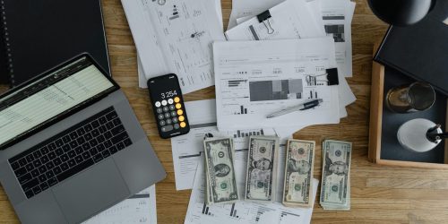 Overhead view of financial documents, cash, and technology on a wooden desk.