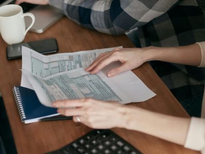 Close-up of hands organizing tax forms on a desk with a calculator, laptop, and notebook.