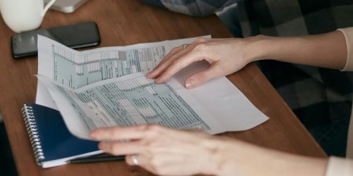 Close-up of hands organizing tax forms on a desk with a calculator, laptop, and notebook.